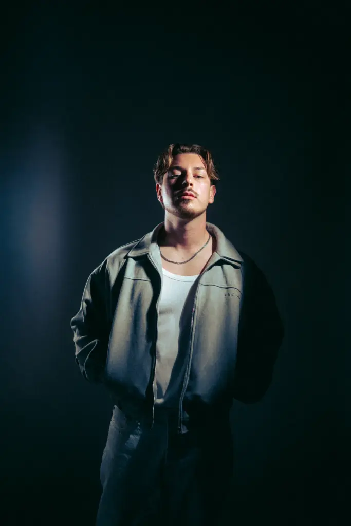 DJ Wamax in heller Jacke vor dunklem Hintergrund