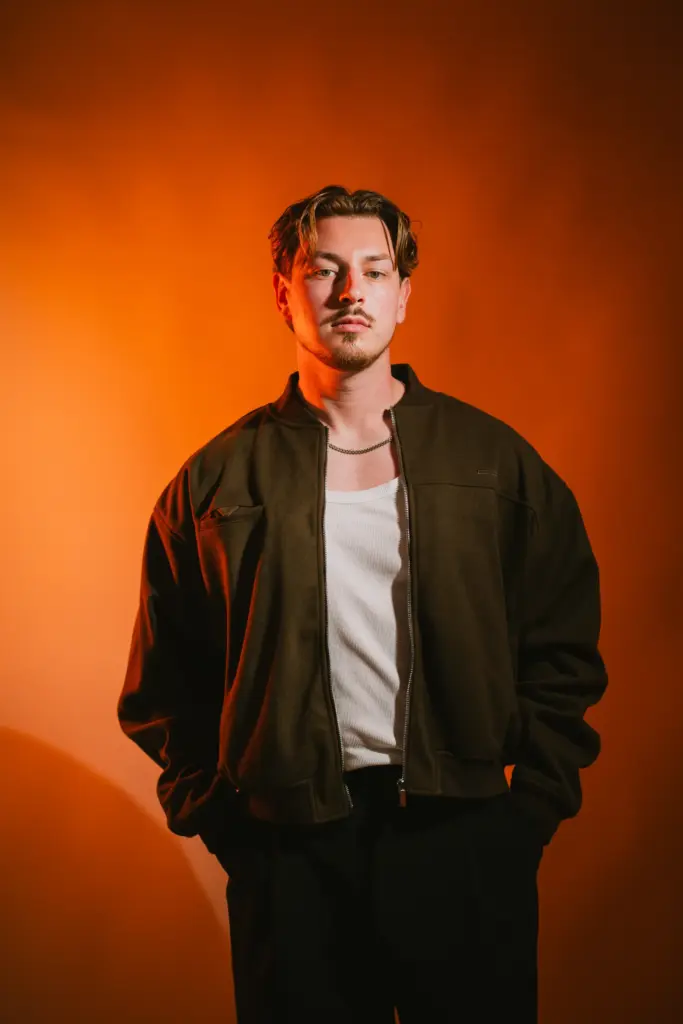 DJ Wamax in dunkler Jacke vor orangem Hintergrund